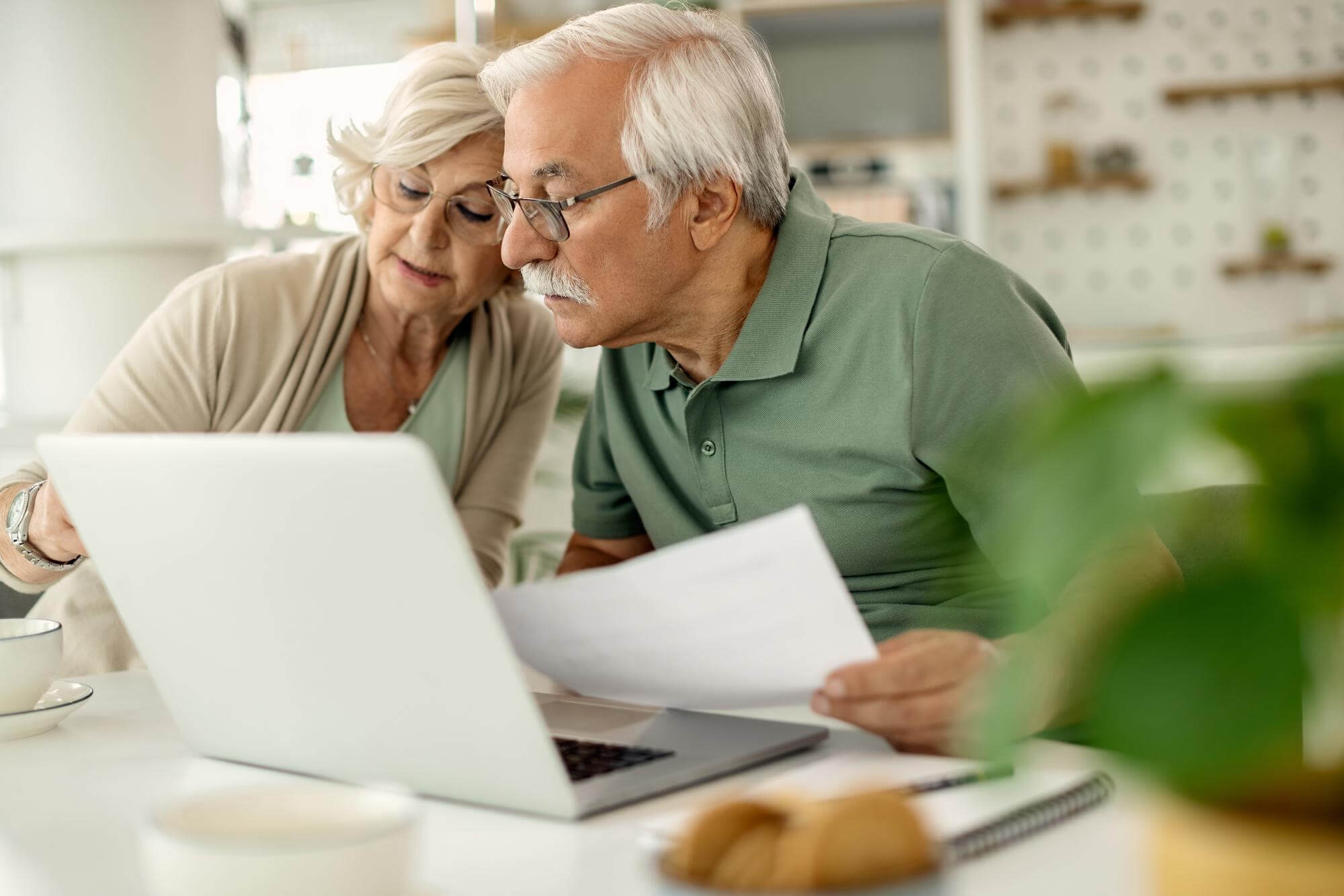 Älteres Ärzte Ehepaar geht in der Küche am Esstisch mithilfe eines Laptops die Informationen zur Energiepreispauschale durch